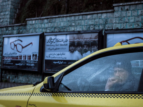 A taxi driver in his car on a rainy day. Behind him a poster of an upcoming performance of Samuel Beckett’s play "Waiting for Godot".© Newsha Tavakolian for the Carmignac Foundation