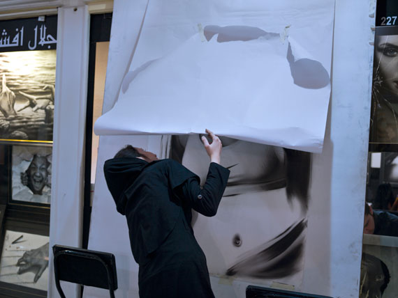 A young woman tries to sneak a peek of a drawing of a woman covered by white paper in Qaem Mall,under Iran’s Islamic laws showing women’s bodies in public is forbidden. © Newsha Tavakolian for the Carmignac Foundation