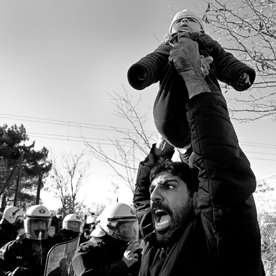 Nikos Pilos: Ein iranischer Migrant demonstriert mit seinem in die Luft gehobenen Kind vor der griechischen Polizei für das Recht, die Grenze zu überqueren.