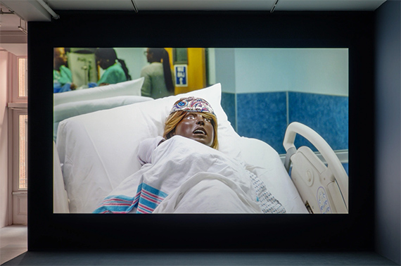 Carolyn Lazard: Two-way. Installation view (Fiction Contract, 2025), Artists Space, 2025. Photo: Greg Carideo. [A darkened gallery space with a video projection centered on a dark blue wall. The projection depicts a close-up of a black birthing mannequin reclined on a hospital bed in a blue patient room. A newborn mannequin swaddled in a hospital receiving blanket lays on the birthing mannequin’s chest.]